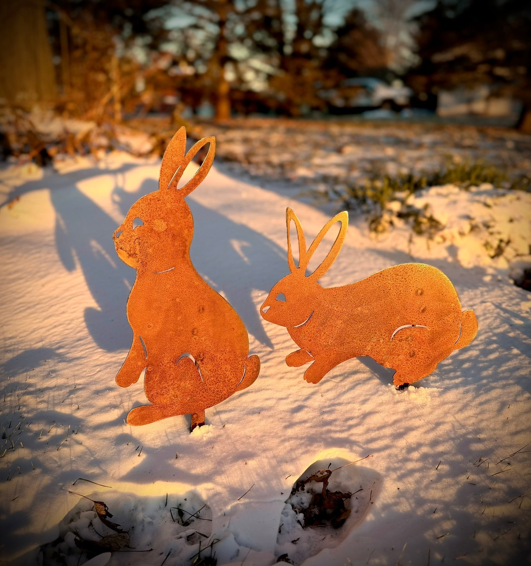 Two rusted metal rabbit sculptures on a snowy ground with a blurred natural background. Henderson Garden Supply