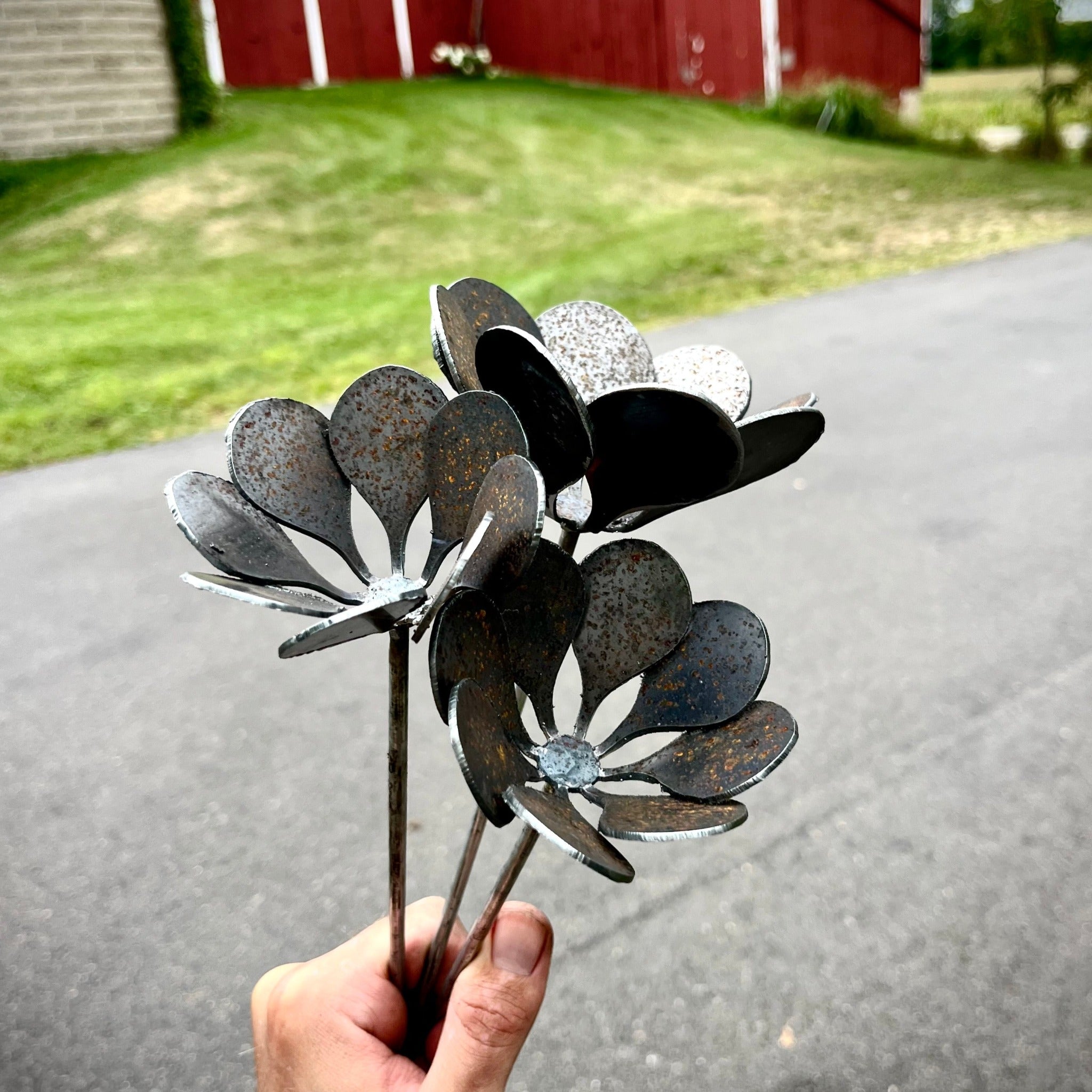 Metal flower sculptures held by a hand with a red barn in the background. Henderson Garden Supply