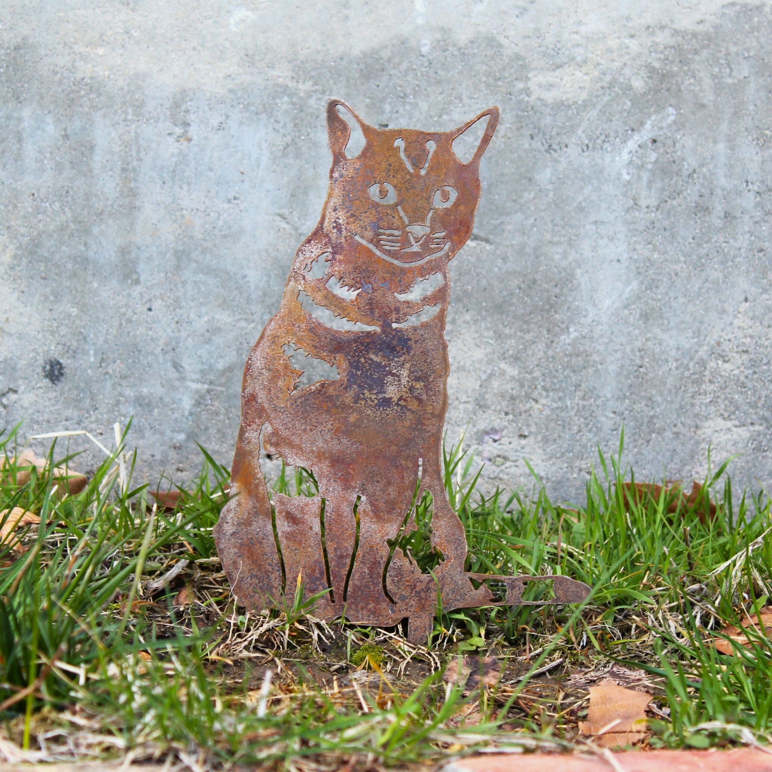Rust-colored metal cat sculpture on grass with a concrete wall background. Henderson Garden Supply
