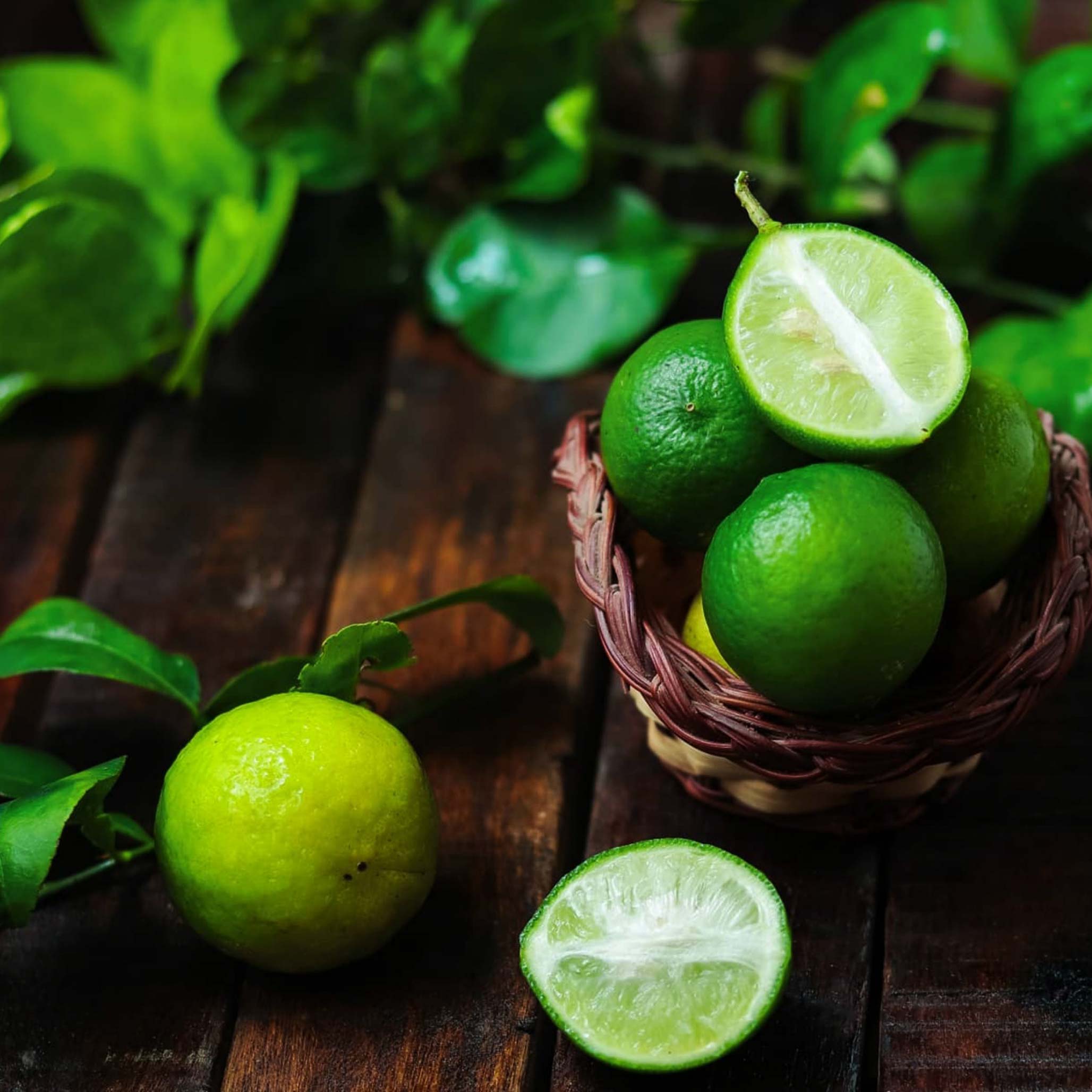 Green limes in a wicker basket on a wooden surface with leaves. Henderson Garden Supply