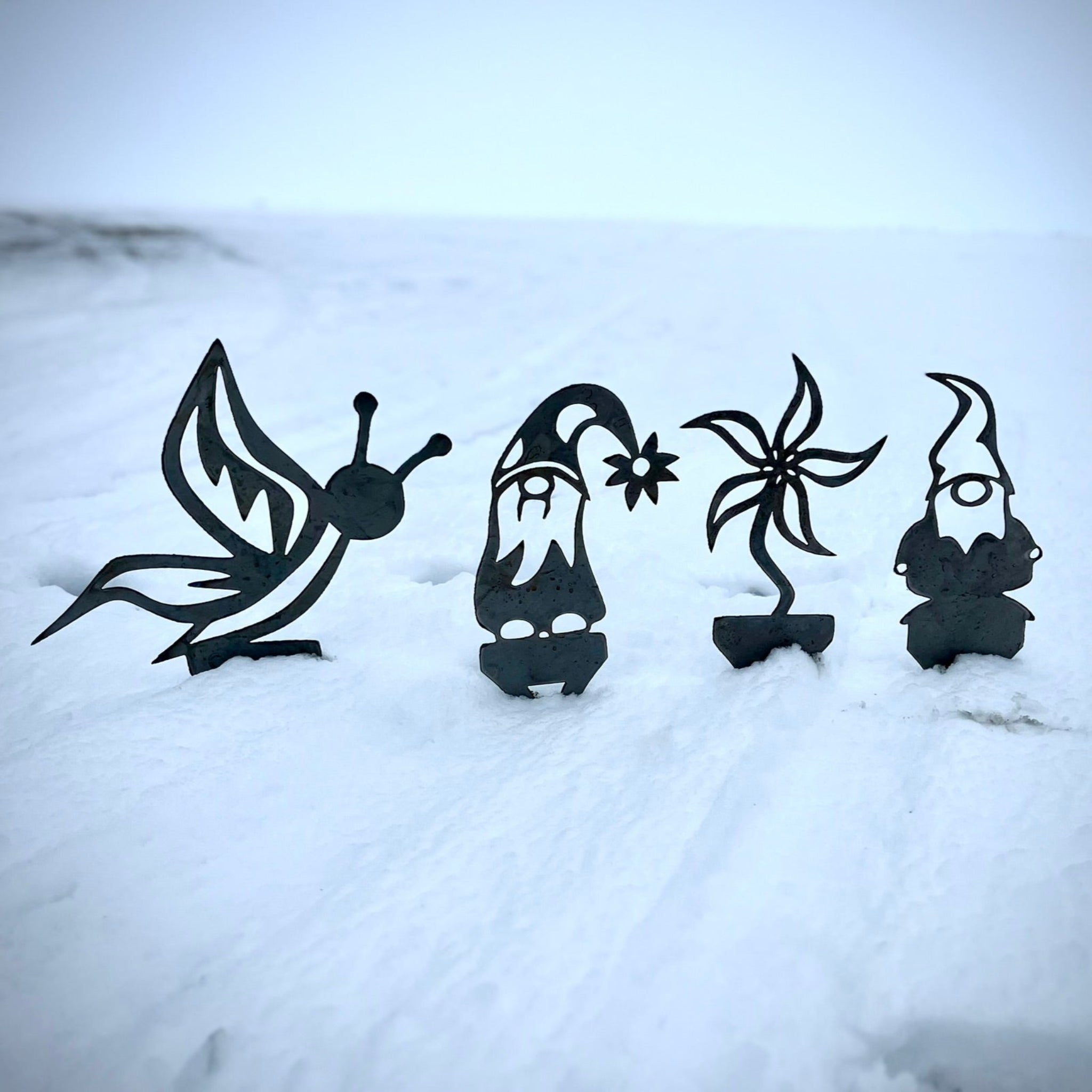 Four black metal garden ornaments shaped like a butterfly, gnome, and two other figures on a snowy background. Henderson Garden Supply