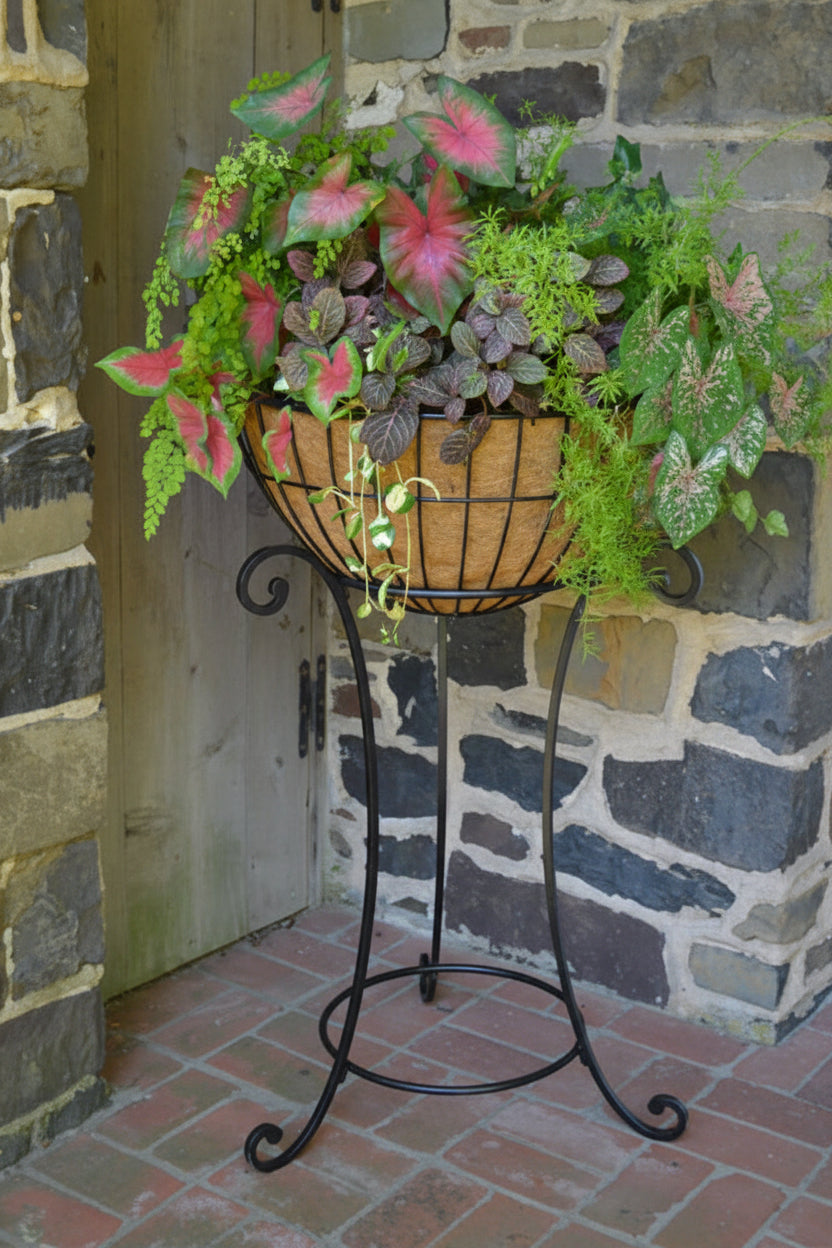 Decorative plant stand with a basket of green and pink plants against a stone wall.  Henderson Garden Supply