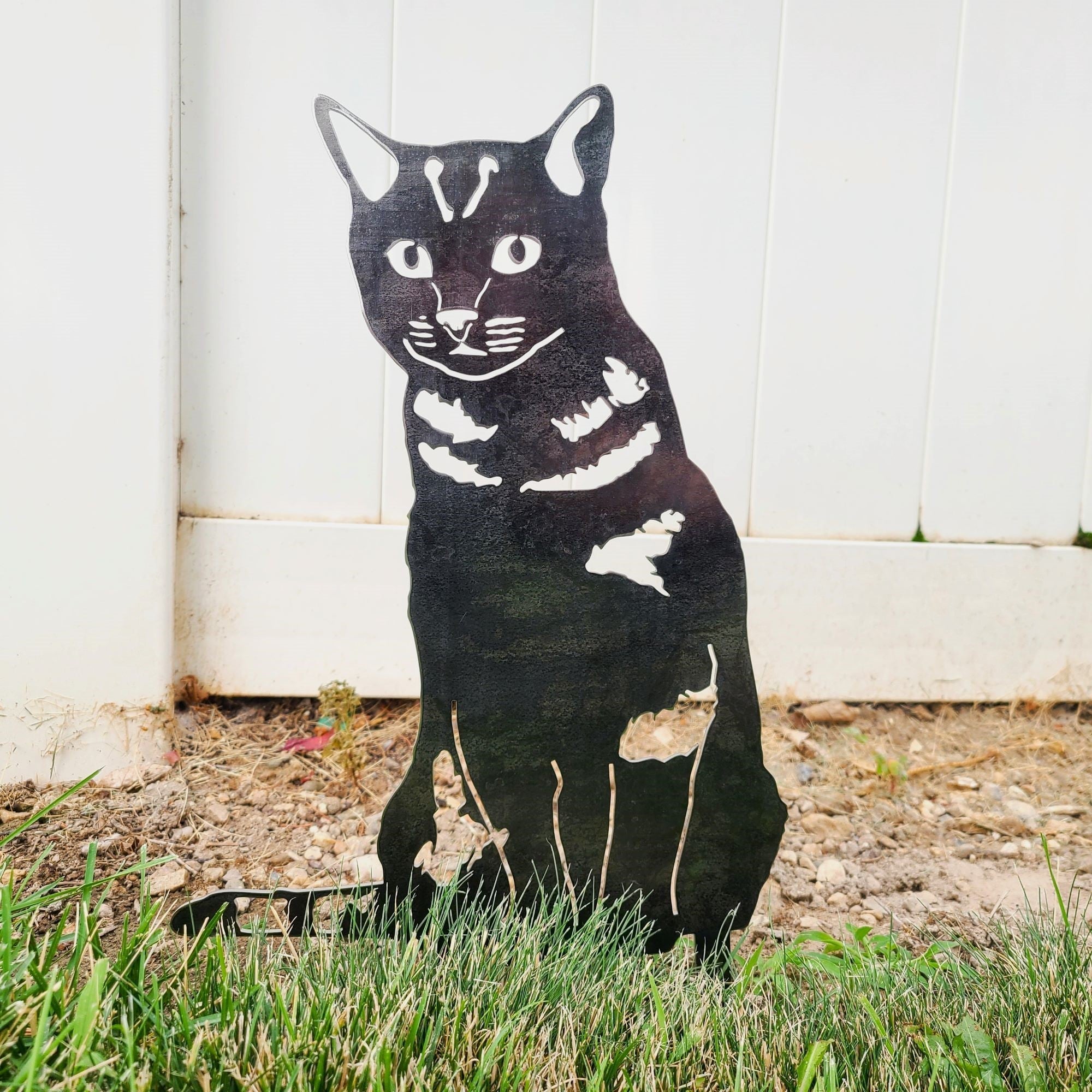 Black cat-shaped decorative object on grass with a white fence background.  Henderson Garden Supply