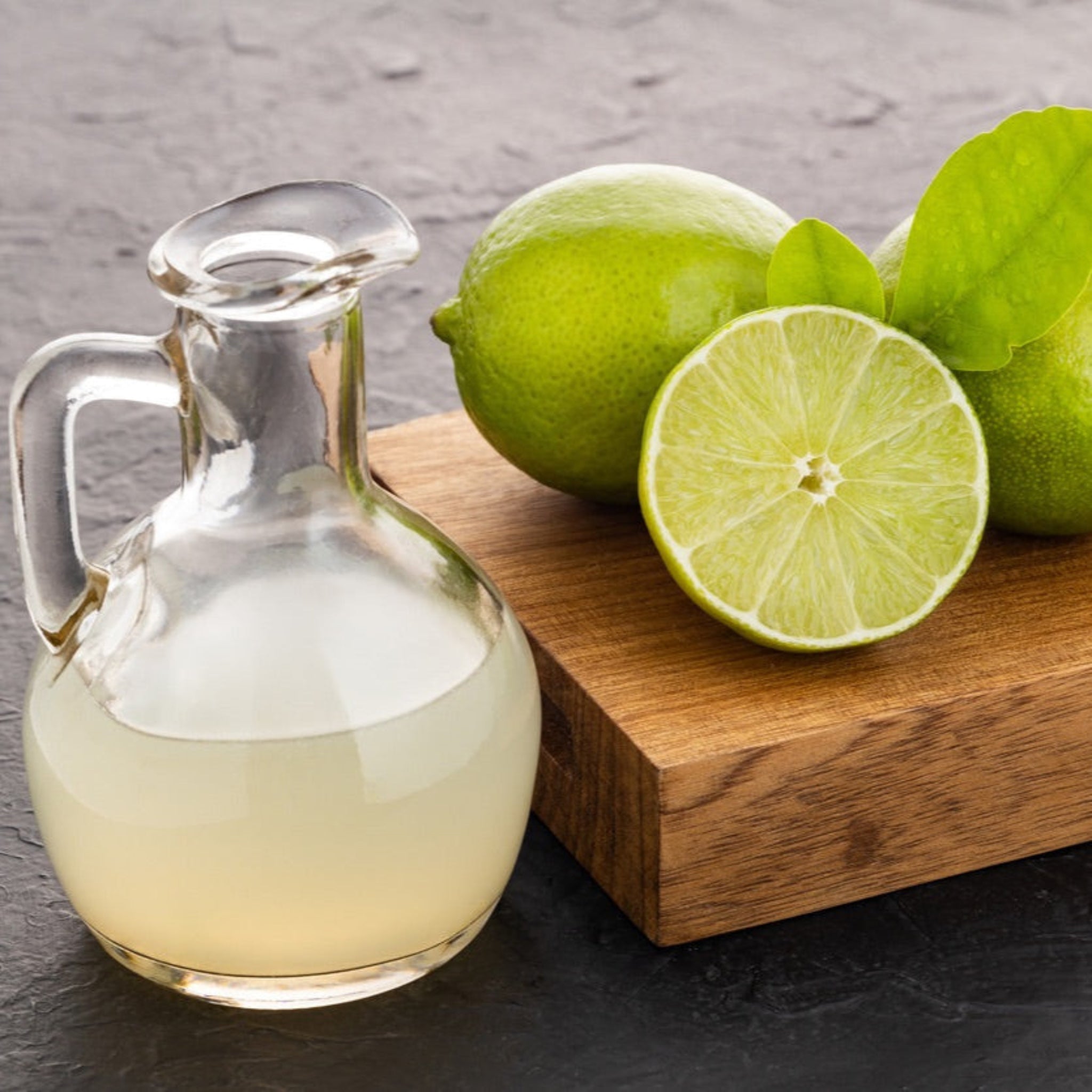 Clear glass container with a liquid next to whole and sliced limes on a wooden board. - Lime Tree - Henderson Garden Supply