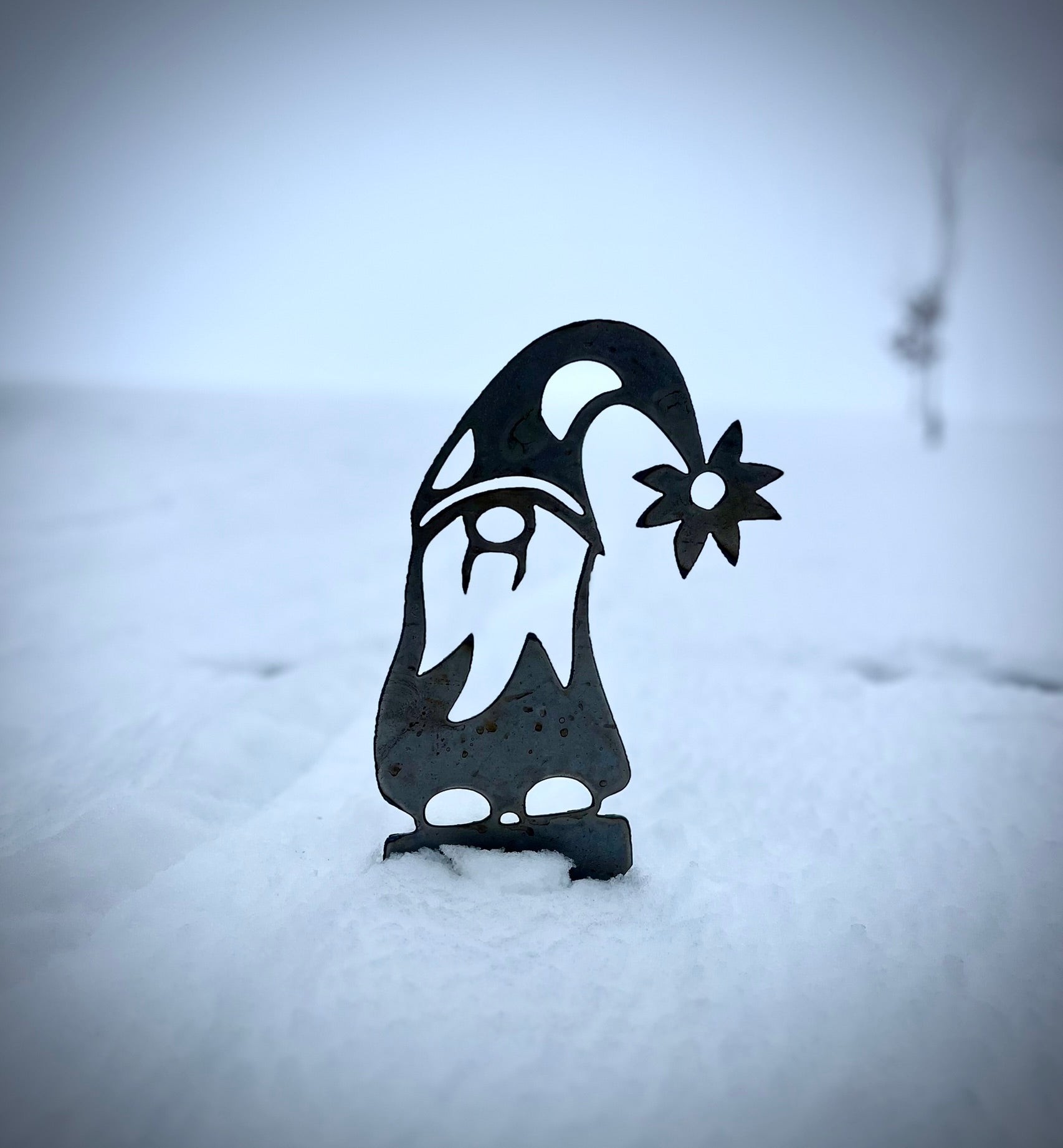 Metal garden gnome sculpture with a flower on a snowy background.  Henderson Garden Supply
